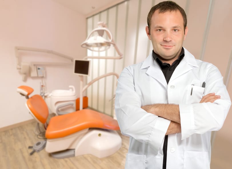 A male dentist standing with his arms crossed and an orange treatment chair in the background