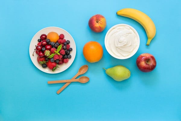 A plate of berries, bowl of yogurt, two spoons and other fruit on a light blue background