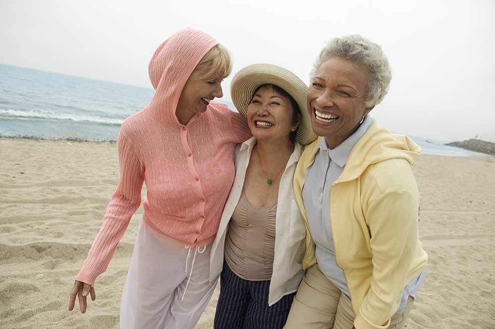 Three smiling older women walking on the beach with their arms around each others shoulders