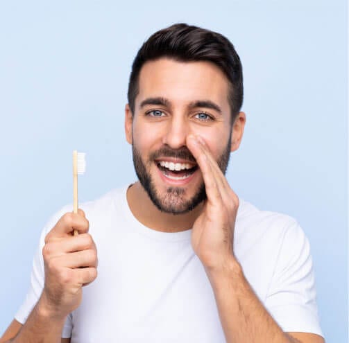 A man with dark hair holding a toothbrush in one hand and holding his other hand up as if whispering
