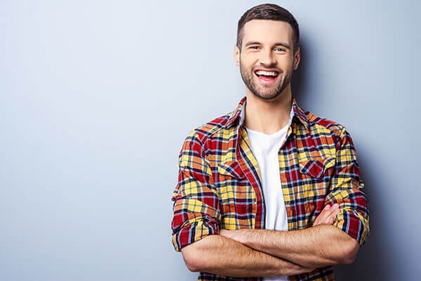 A smiling man in a red, yellow, and orange plaid shirt standing against a light blue background
