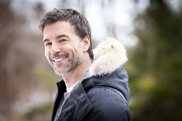 A smiling man wearing a fur lined winter coat with trees blurred in the background